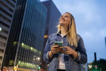 Young woman using a smartphone at night time with city view landscape in the background. High quality photo