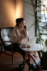 Distance learning. Asian female student sitting in cafe, looking at phone and writing in writing notebook.