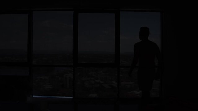 A shirtless, young male with medium length hair walks into frame and slowly opens a single window blind to reveal a the beautiful Gold Coast City view below which he stands and observes for moments