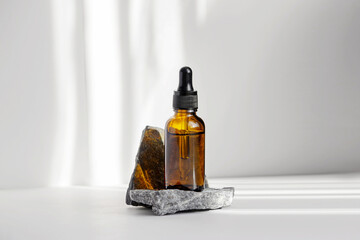 Serum in glass bottle with dropper lid on stand made of natural granite stone. Essential oil for care of women's skin on white background, in rays of sunlight. Space for copying, glare on object
