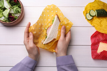Woman packing sandwich into beeswax food wrap at white wooden table, top view