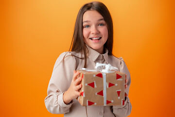 Cute teen girl holding a Christmas gift box against orange background.