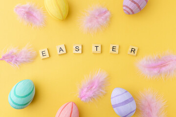 Easter composition with an inscription and colorful eggs and feathers on a yellow background, top view. Preparation for the holiday.