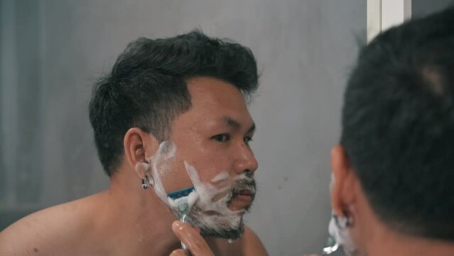 Adult man shaving with foam and manual razer.Mirror reflection young handsome smiling man touching beard, grooming in bathroom. Head shot close up happy asian man guy doing morning beauty routine.