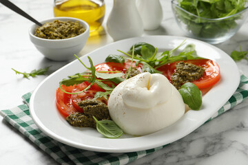 Delicious burrata salad with tomatoes, arugula and pesto sauce served on white marble table, closeup