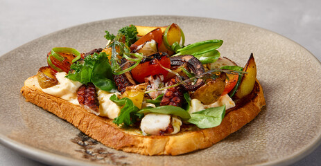 Bruschetta with octopus, vegetables and sauce on gray ceramic plate. Toast with octopus in contemporary style. Chef concept bruschetta in modern ceramic dishware. Handmade dinner plate.