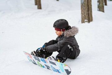 Fototapeta na wymiar little boy sitting on snow putting his feet in snowboard bindings adjusting straps