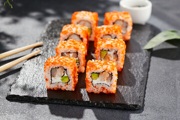 Maki sushi on dark stone table. California maki with tobiko. Sushi roll with cheese,shrimp, avocado inside, maguro outside. Style concept japanese menu with black background, leaves and hard shadow.