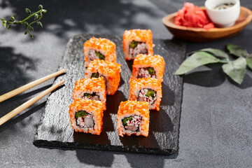 Maki sushi on dark stone table. California maki with tobiko. Sushi roll with crab,shrimp, cucumber inside, maguro outside. Style concept japanese menu with black background, leaves and hard shadow.