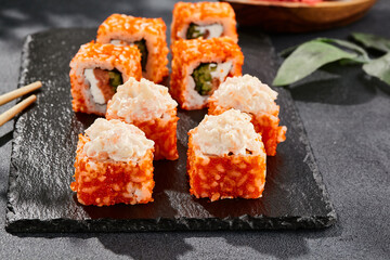 Maki sushi on dark stone table. California maki with tobiko. Sushi roll with salmon, cucumber inside, maguro, crab outside. Style concept japanese menu with black background, leaves and hard shadow.