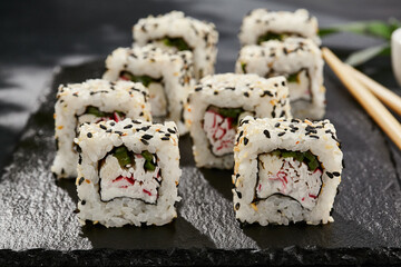 Maki sushi on dark slate. California maki with sesame. Sushi roll with crab, cucumber inside, sesame outside. Style concept japanese menu with black background, leaves and hard shadow.