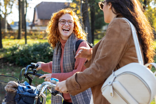 Two young woman with bikes walk and talk and smile in public park