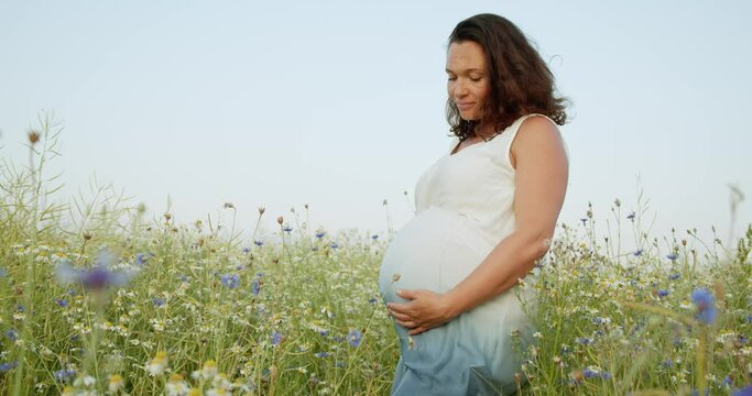 Pregnant woman with a big belly feeling happy outdoors. Adult expecting mother stroking belly on nature.  Middle age white pregnant woman touching her belly while sunset. Woman pregnancy concept.