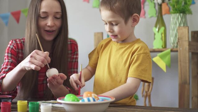 Mom and a child are painting eggs together in a room decorated for the holiday and having fun. Concept of preparing for Easter, spring mood and happy family