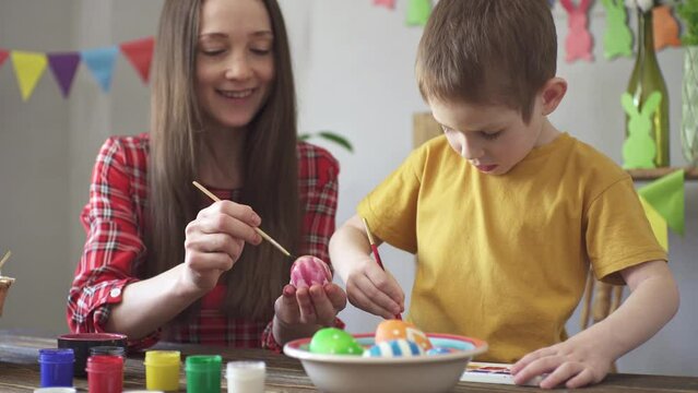 Mom and a child are painting eggs together in a room decorated for the holiday and having fun. Concept of preparing for Easter, spring mood and happy family