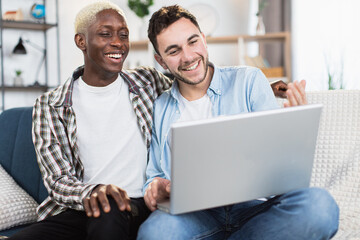 Sweet couple of multiracial gays smiling cheerfully while looking on laptop screen. Two people in...