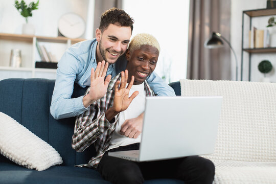 Happy and beautiful gay couple smiling and waving during video call on modern laptop. Multiracial men having online conversation while sitting on cozy sofa.