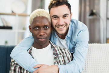 Fototapeta premium Portrait of positive men in relationship embracing on couch while smiling and looking at camera. Two multicultural gays enjoying time spending together.