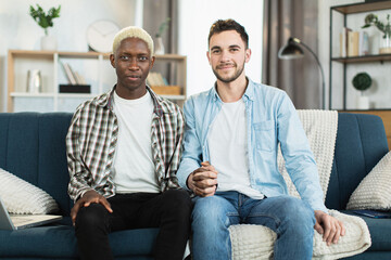 Two multicultural men in relations holding hands while sitting together on comfy couch. Freedom and closeness of lgbtq people.
