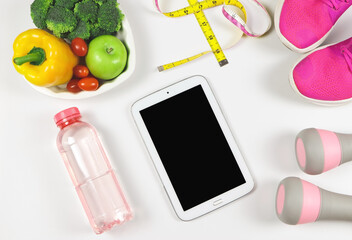  flat lay of black blank screen computer tablet , vegetables and fruit in heart shape plate, bottle of water, measuring tape,  pink dumbbells and pink sneakers on white background.