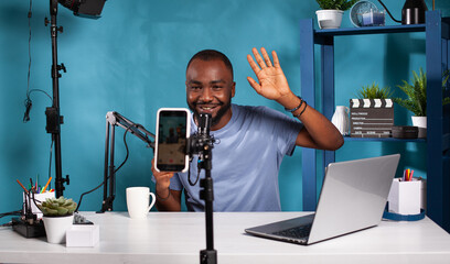Fototapeta na wymiar Content creator doing greeting hand gesture looking at live video podcast setup sitting at desk with professional microphone. Smiling vlogger waving hello in front of recording smartphone.