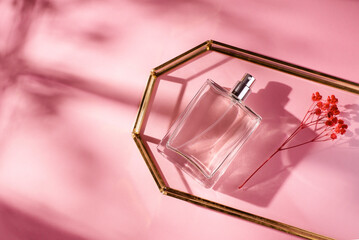 Transparent bottle of perfume in glass tray on a pink background. Fragrance presentation with daylight with red flowers. Trending concept in natural materials with window shadow. Women's essence.