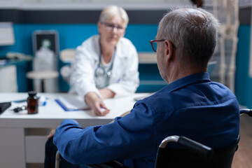 Fototapeta premium Close up of patient with disability attending checkup appointment with doctor at clinic. Senior man with disease sitting in wheelchair while meeting with physician to help with illness.