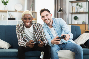 Positive couple of two gays holding console playing games while sitting together on couch. Leisure time with fun of loving people.