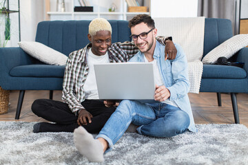 Happy multiracial couple of two gays sitting on floor at living room and using modern laptop. People in love scrolling social networks during free time.