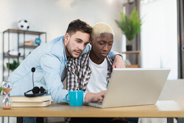 African american man sitting at desk and typing on laptop while his caucasian boyfriend standing near and embracing him. Concept of sexual orientation, technology and lifestyles.