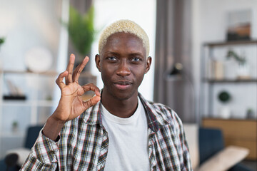 Portrait of african man with blond hair and blue eyes gesturing sign OK with fingers while sitting...