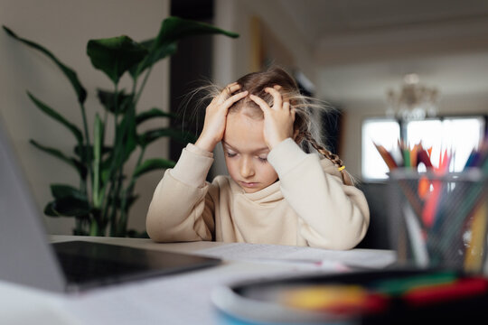 Tired Caucasian kid studying online at home and using laptop. Distance or remote learning for child. Pretty stylish schoolgirl studying homework math during coronavirus