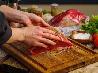 The chef's hands are seasoning a large piece of farm meat on a wooden cutting board. Cooking meat dishes in a restaurant and home kitchen, cookbook, culinary blog, advertising.