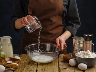 The chef's hands are preparing the dough in a glass bowl on a wooden table on a dark background. Ingredients for making pizza, bread, buns, pies, pasta, confectionery. Cookbook, food blog.