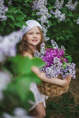 A blonde European girl in a knitted white beret holds a basket of lilac flowers standing in a lilac garden