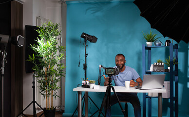 Wide view of vlogging studio setup with smiling vlogger interacting with audience sitting at desk holding cup. Content creator talking to followers in in front of filming video camera on tripod.