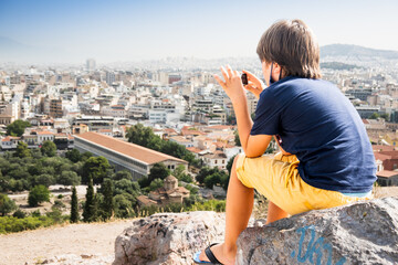 Rear view of boy sitting on stone and photographing cityscape