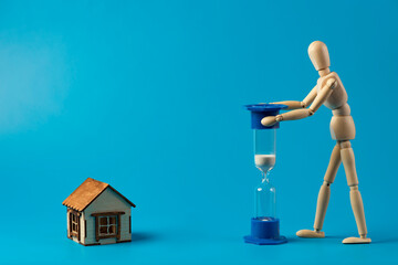 Wooden man hourglass and small house on a blue background. Real estate purchase, mortgage and real estate investment concept. Copy space.