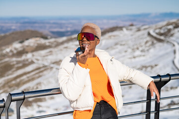 A African American woman having phone conversation leaning against fence in snowy mountain during winter