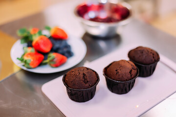 three chocolate muffins on a white cutting board, in the background a plate with berries for decorating cupcakes