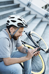 Young bearded cyclist sitting on stairs and looking at phone.