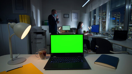 Laptop with green screen on desk and employees in safety mask working on background