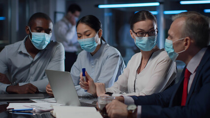 Diverse coworkers in safety mask working on laptop together in office