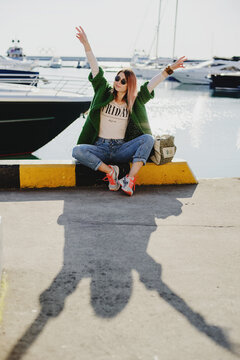 a young girl in jeans, a cardigan and sunglasses seets laughing by the sea against the background  yachts