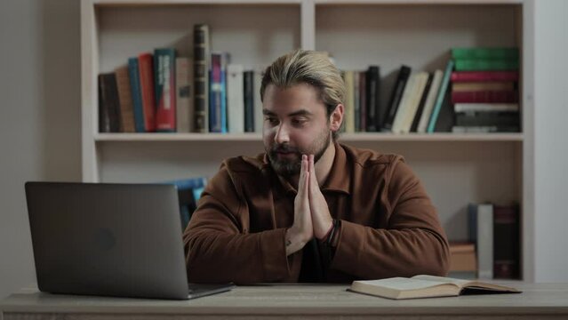 Hopeful caucasian man sitting at desk in hands folded in prayer gesture and looking on computer screen with excitement. Asking for success and luck.