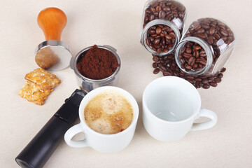 Cup of espresso, crackers, holder with ground coffee, tamper and cans of coffee beans on table.