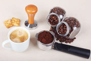 Cup of espresso, crackers, holder with ground coffee, tamper and coffee beans on table.