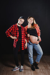 A pregnant girl in jeans and a black T-shirt with teen girl. Photo shoot in the studio on a black background