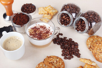 Breakfast with cup of espresso, cappuccino with chocolate crumbs, cookies, crackers, holder with ground coffee, tamper and coffee beans on table.