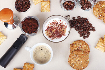 Cup of coffee, cappuccino with chocolate crumbs, crackers, cookies, holder with ground coffee, tamper and coffee beans on table. View from above.